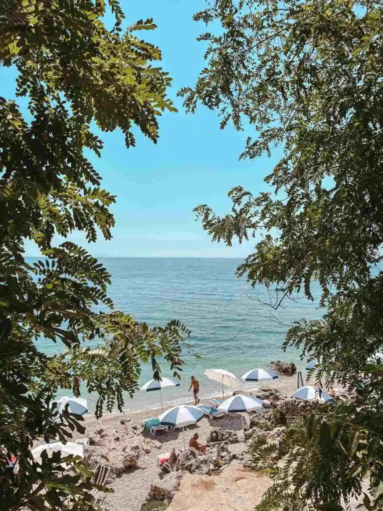 Crystal-clear turquoise water laps against the pebbled shore of Dhermi Beach in Albania, framed by rocky cliffs and lush greenery. Sunbeds and umbrellas line the coast under a bright summer sky, creating a relaxed Mediterranean vibe.