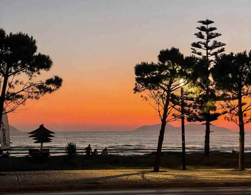 A golden sunset casts a warm glow over the Lungomare promenade in Vlorë, Albania, with palm trees silhouetted against the orange-pink sky and the calm sea shimmering in the fading light. Locals and tourists stroll along the waterfront enjoying the view.
