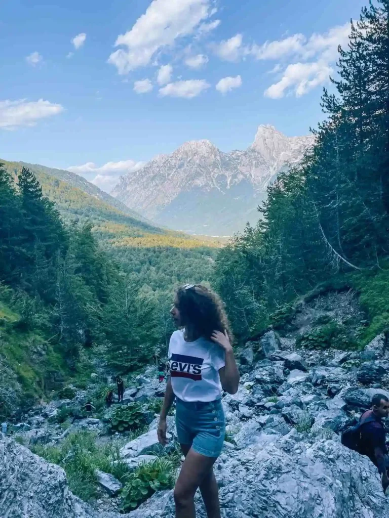 A breathtaking view of the Thethi Mountains in Shkoder, Albania, with rugged alpine peaks rising above lush green valleys and a clear blue sky. Traditional stone houses and winding trails dot the remote, untouched landscape.