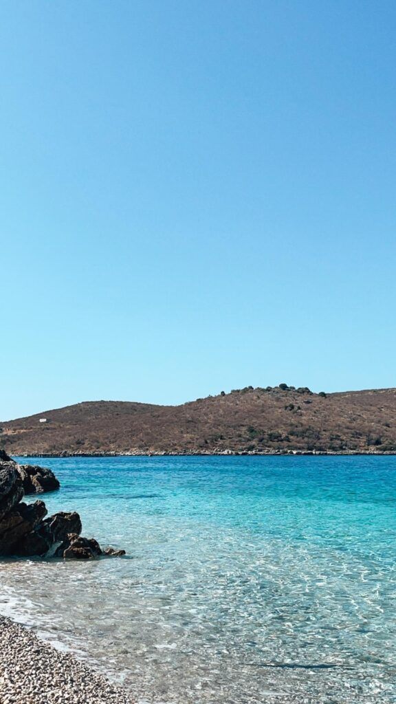 A peaceful beach at Porto Palermo with deep blue water and rugged hills in the background. The secluded yet serene setting highlights the slower pace of travel that can make Albania feel safe and manageable when traveling alone.
