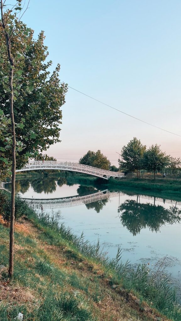 Riverside scene along the Drini River lined with numerous cafés and outdoor seating. People relax at tables under umbrellas as sunlight glints on the flowing water, making it a lively spot for digital nomads in Albania.