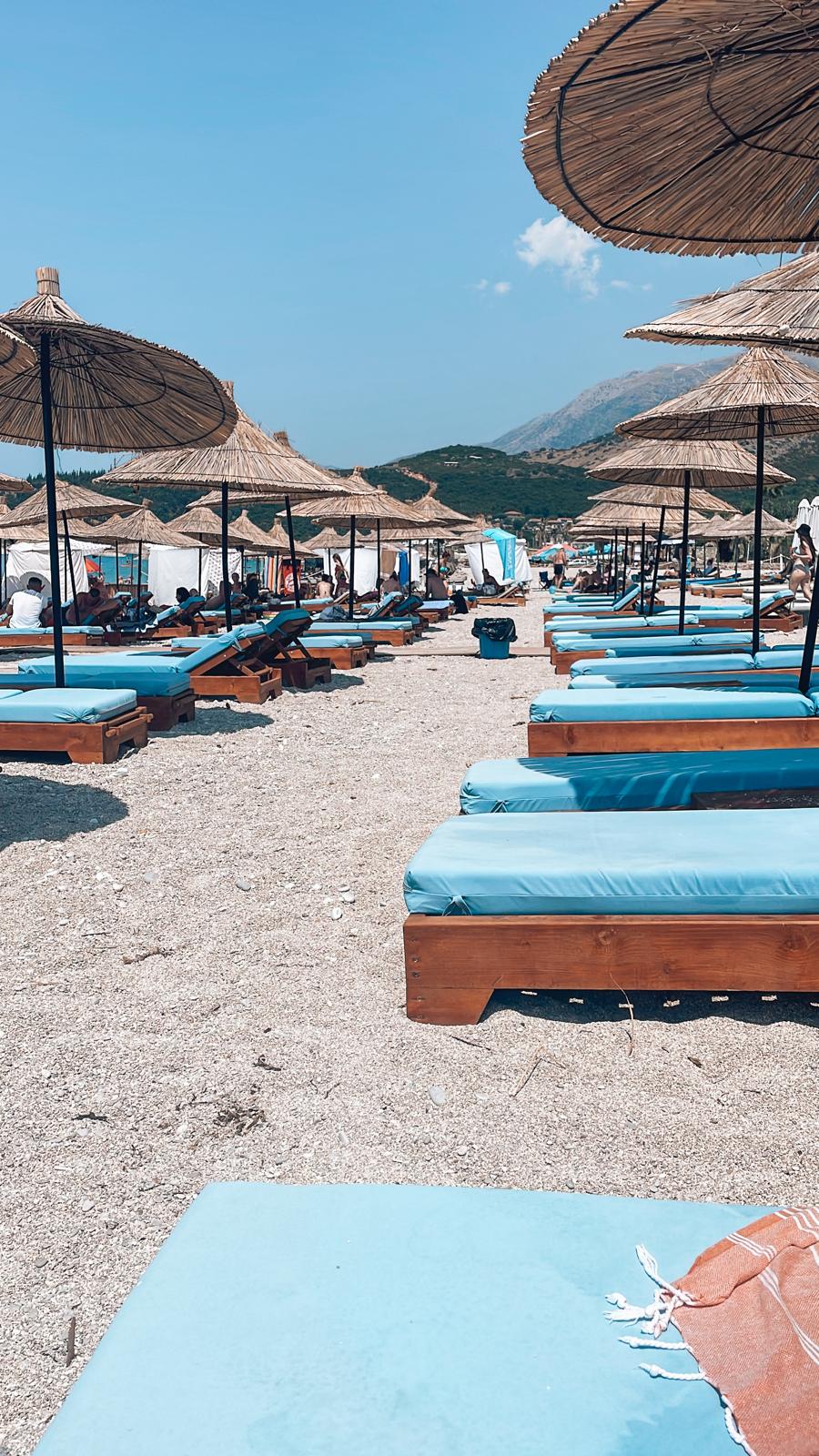 A wide view of Drymades beach with clear turquoise water meeting a pebbled and sandy shoreline under an open sky. The scene captures a more natural and spacious coastal feel often contrasted in the drymades vs Dhermi comparison.