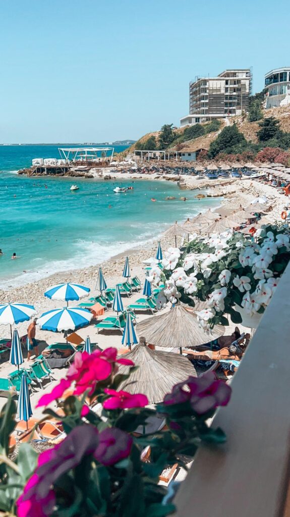 Sandy shoreline of Orikumi Beach with gentle waves and sparse beachgoers under a clear sky. Quiet coastal stop along an Albania road trip for seaside relaxation.