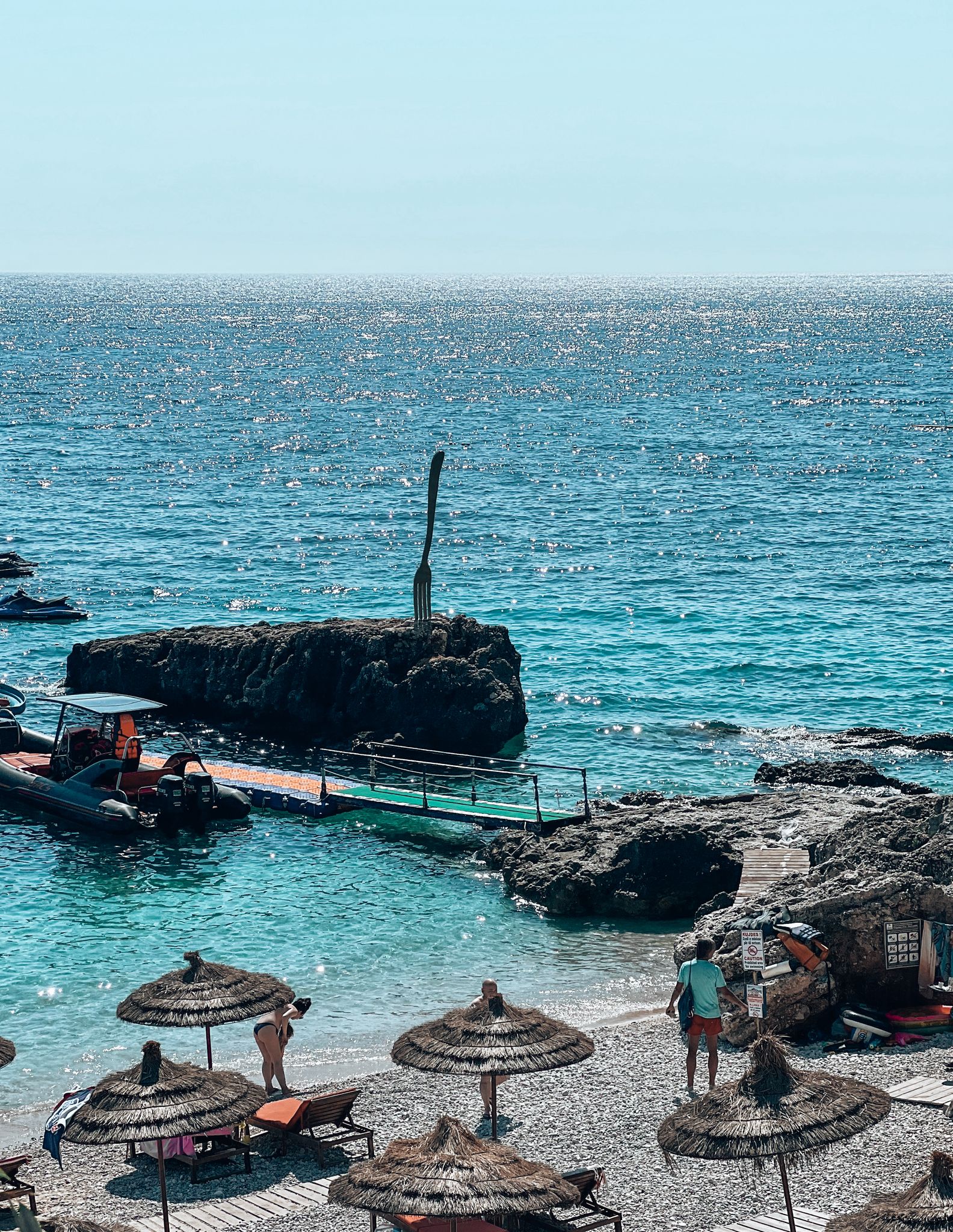 Deep blue sea along the coast of Dhërmi in Albania with gentle waves meeting a pebbled shoreline and a hillside village in the background. This scenic coastal view highlights the best area to visit in Albania while hinting at common travel mistakes like underestimating distances between seaside towns.