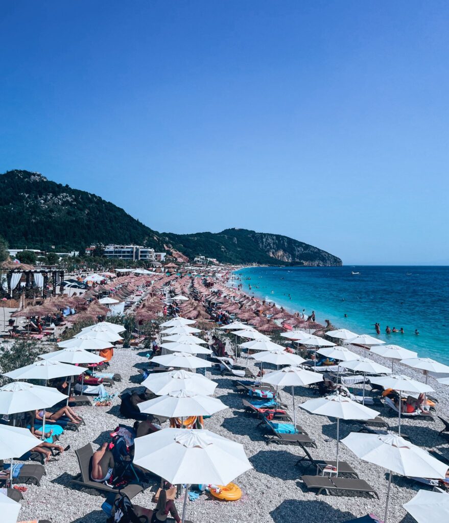 Wide view of Dhermiu Beach with soft white sand and clear turquoise water stretching into the horizon. A few sun loungers and umbrellas dot the shore, showing a relaxing destination for traveling Albania Without a Car.