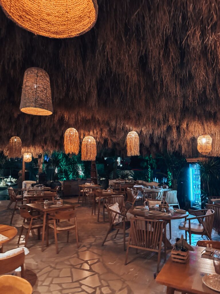 Cozy interior of a beach bar with wooden tables, hanging lights, and colorful décor. Large windows open to the seaside, creating a casual spot to relax while exploring Albania Without a Car.