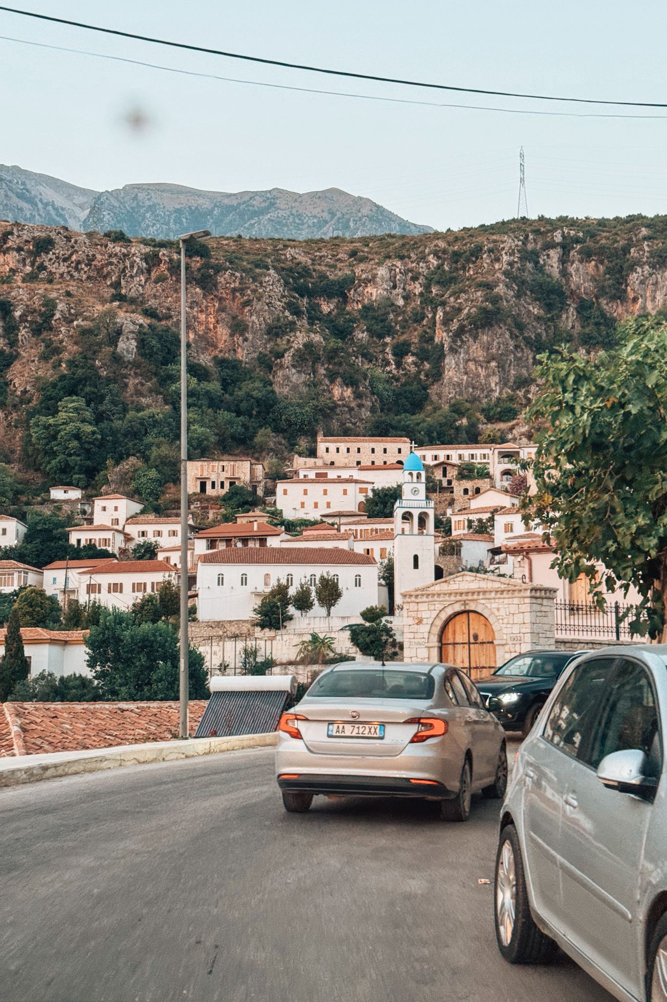 A coastal village in Dhermi with clustered buildings set against hillside terrain and views toward the Ionian Sea. The scene captures the lived-in seaside charm of the area often referenced in the drymades vs Dhermi comparison.