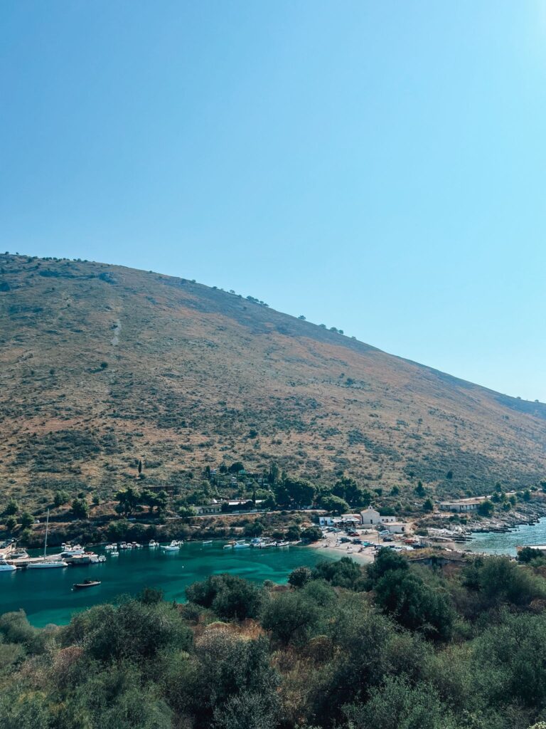 Potami Beach with clear blue water meeting a wide stretch of pebbled shore backed by rocky cliffs and greenery. The natural coastal scenery highlights a scenic stop while backpacking Albania along the Ionian coast.