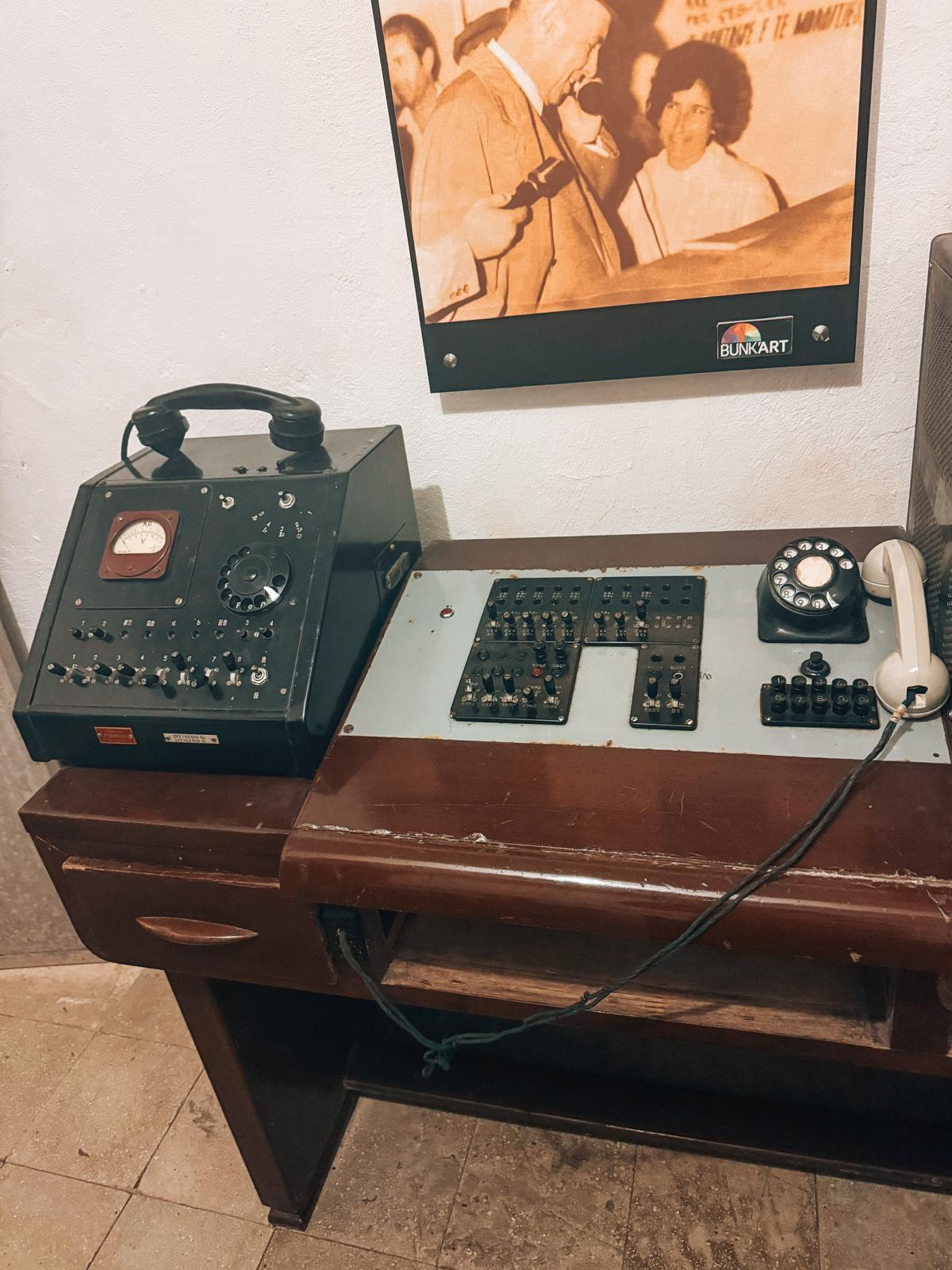 An old telephone or fax machine sits on a worn surface with visible signs of age and use, representing outdated communication technology from the communist era. The device helps illustrate the historical setting encountered when visiting a bunker in Albania.