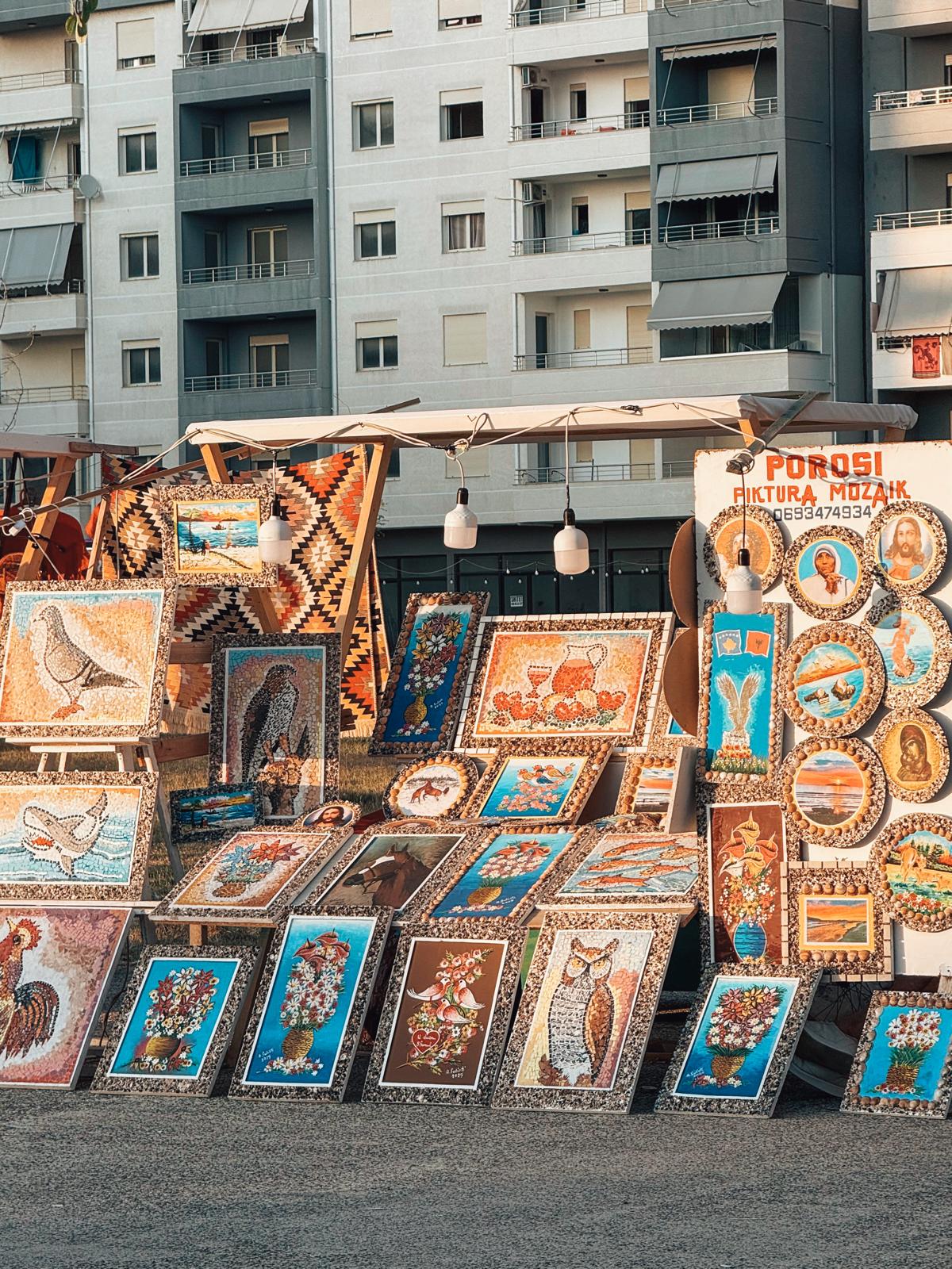 Street-side handmade carpet shop in Tirana displaying colorful woven rugs hanging vertically outside the storefront under warm evening lighting. The scene reflects a traditional craft-focused side of Tirana nightlife with artisan goods showcased along a lively city street.