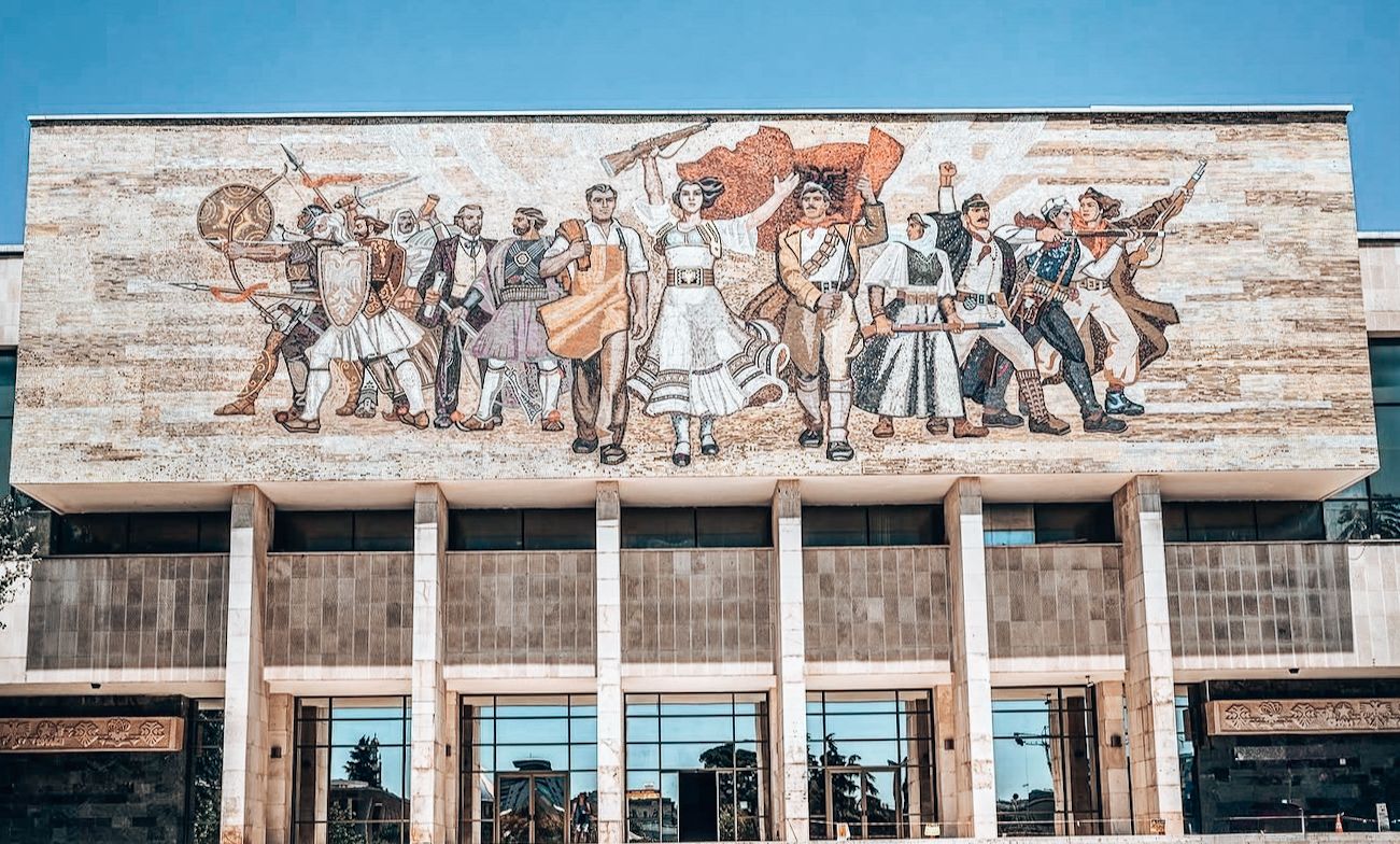 The exterior of the National Historical Museum shows its large mosaic-covered facade, viewed from Skanderbeg Square in the heart of the city. The wide square in front feels open and lively, with urban architecture framing the scene. A key stop for 48h in Albania, it highlights the city’s history right at its central landmark.