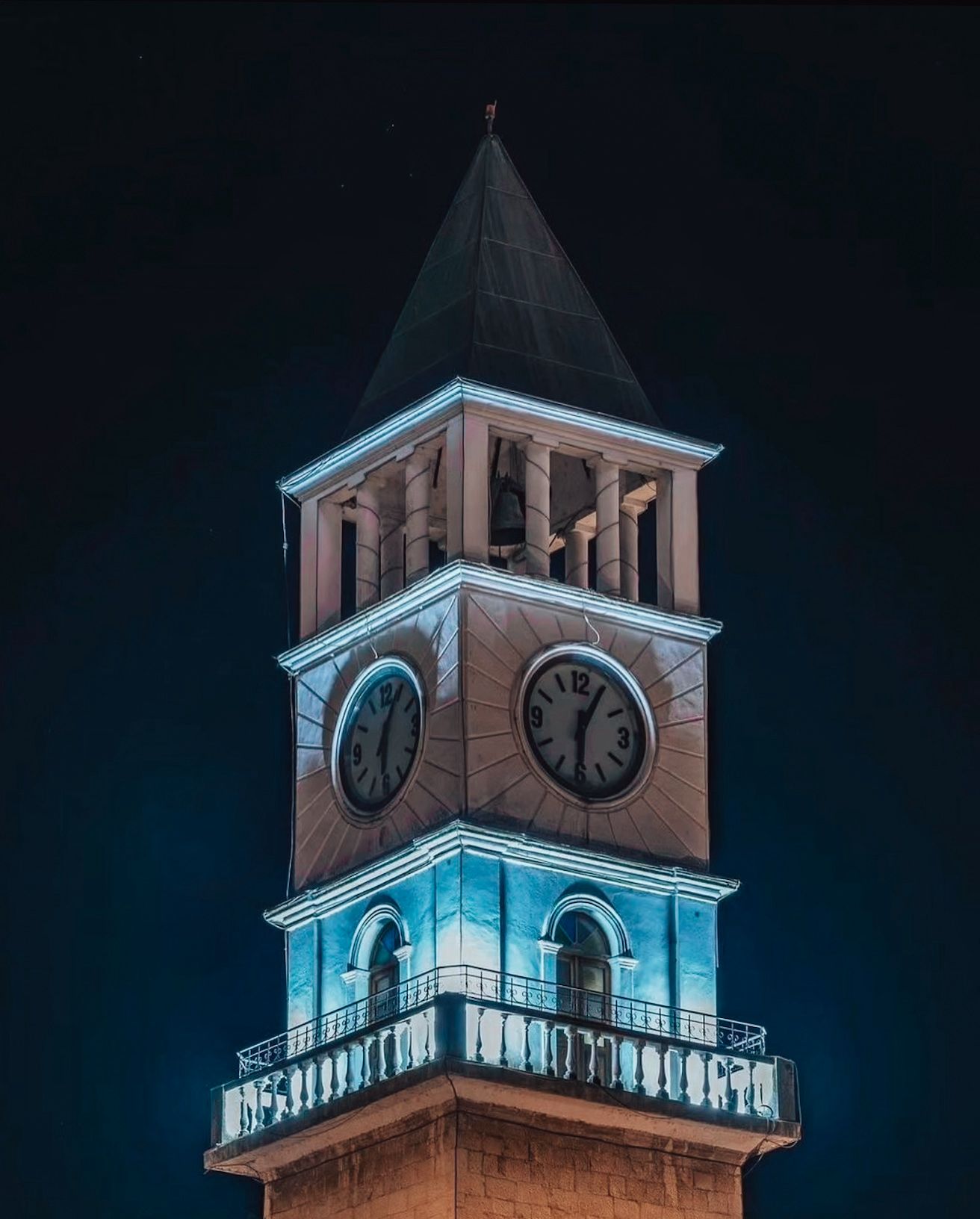 Tirana Clock Tower illuminated at night with warm golden lighting highlighting its historic stone structure against a dark sky. The surrounding square is softly lit, reflecting a calm yet atmospheric Tirana nightlife mood in the city center.
