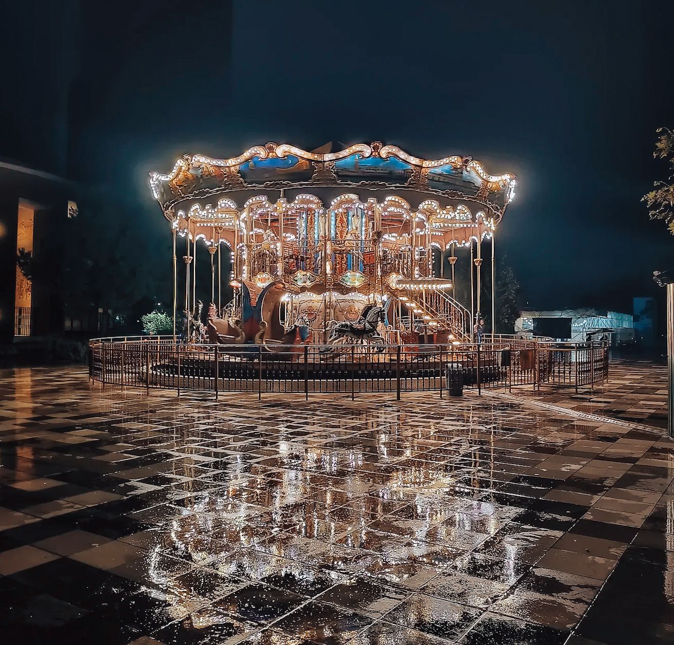 Skanderbeg Square in Tirana at night illuminated with warm city lights, featuring a brightly lit carousel in the center with motion blur suggesting movement. The scene captures a lively Tirana nightlife atmosphere with surrounding buildings glowing against the dark sky.