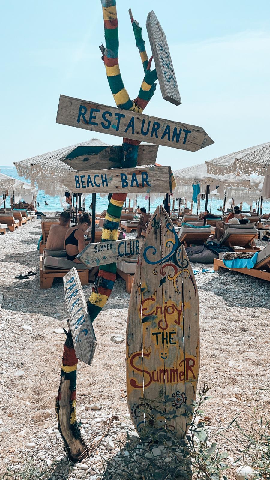Alt text: Stylish beach decor at a seaside restaurant on the Albanian Riviera with the restaurant name written on a decorative surfboard surrounded by lounge seating and coastal details.