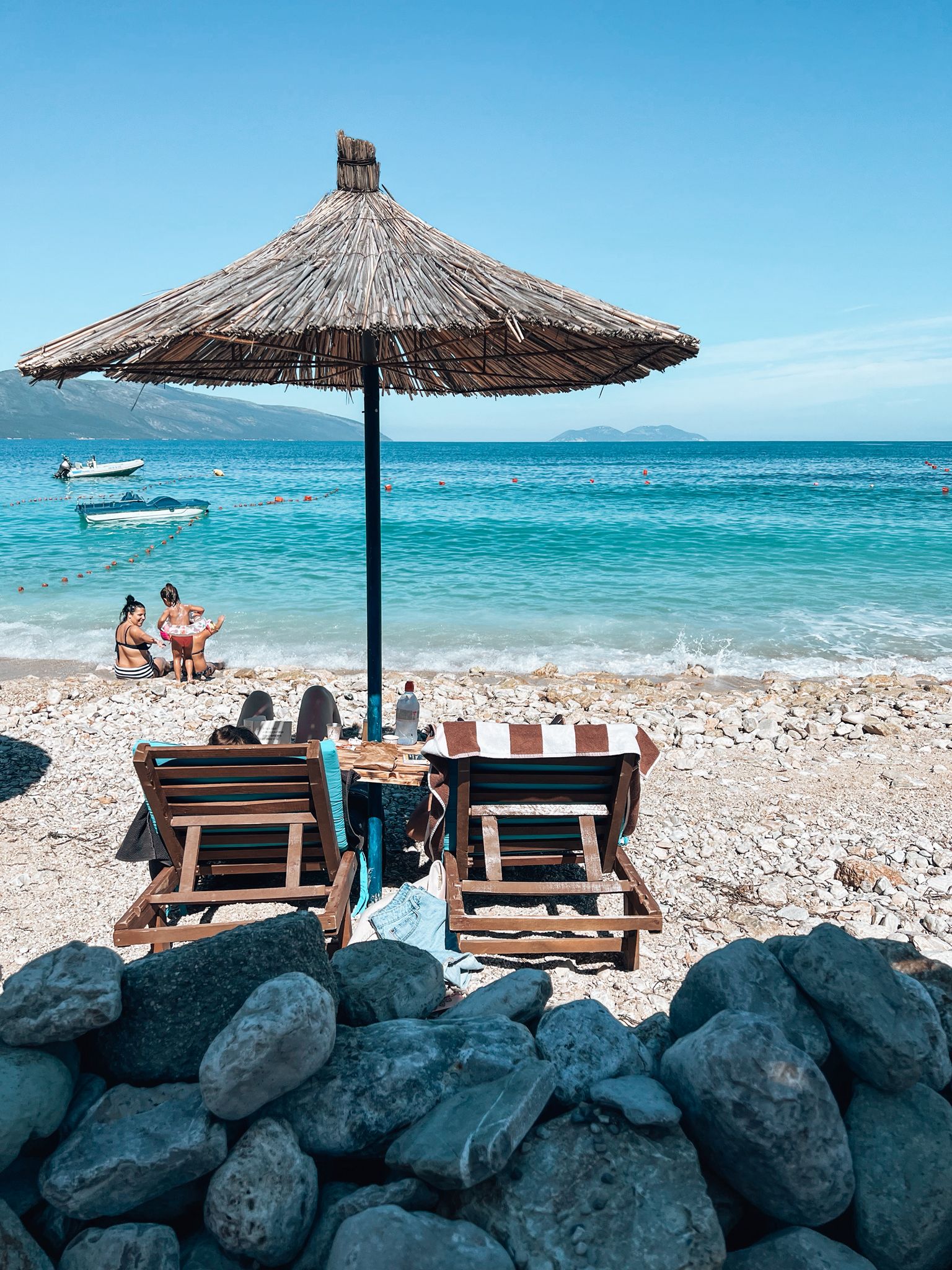 Alt text: Pebbled shoreline and calm turquoise water at Orikumi Beach near Vlore on the Albanian Riviera with mountains visible in the distance.