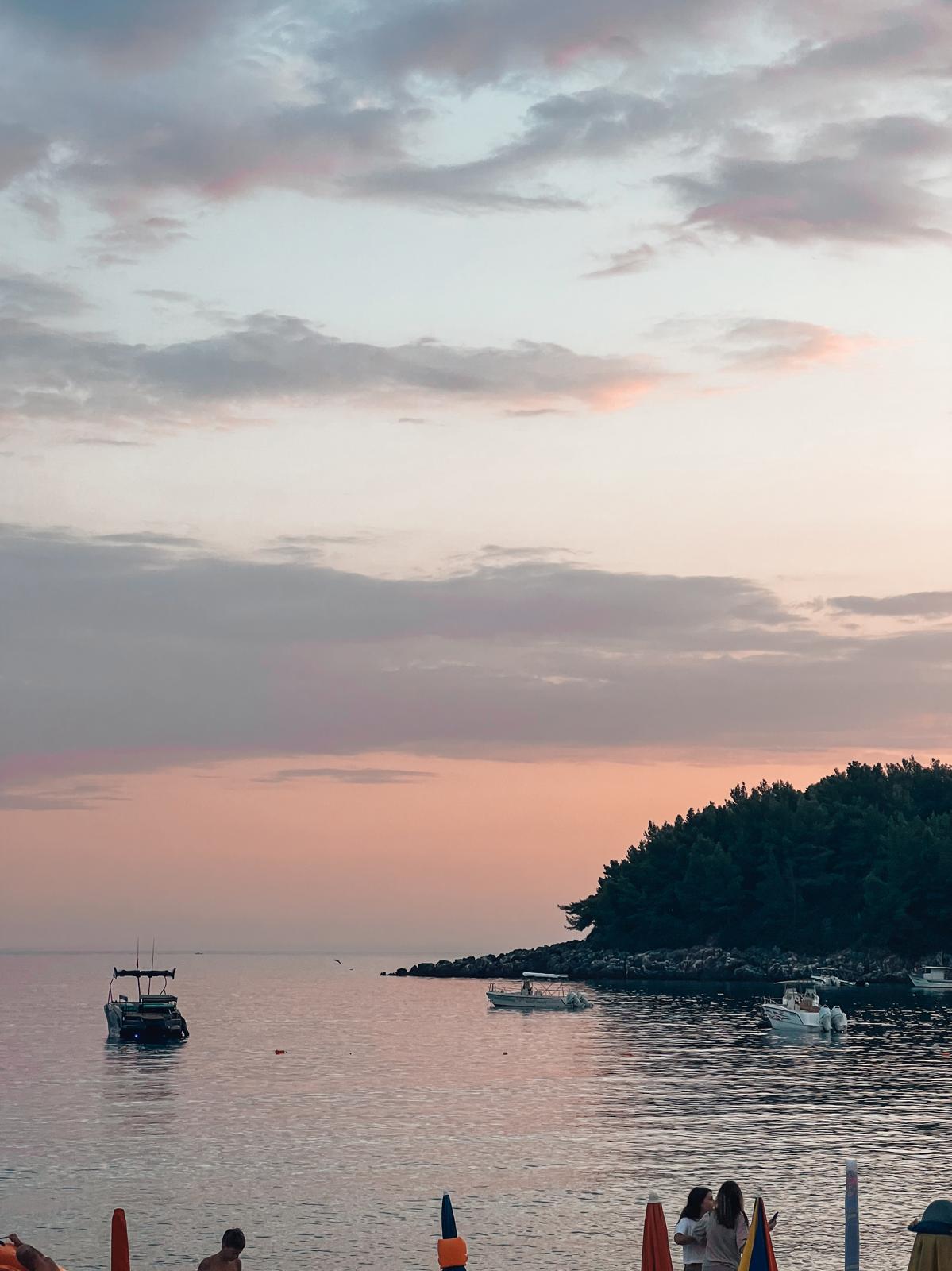 Golden sunset over Spile Beach on the Albanian Riviera with calm waves, a rocky shoreline, and warm light reflecting across the sea.