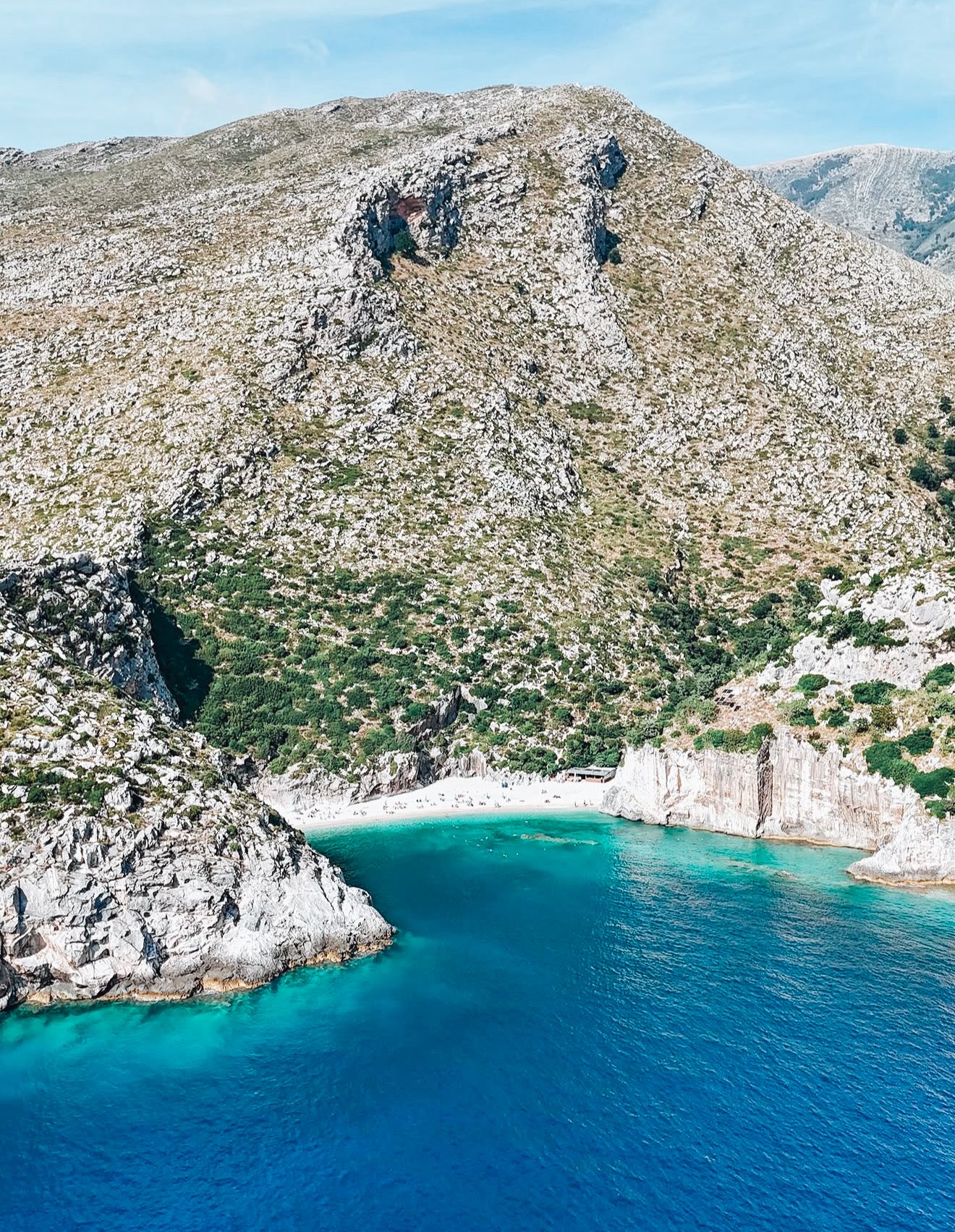 Alt text: Aerial view of Grama Bay in Albania with turquoise water curving along a rocky coastline and small boats floating near the beach. The dramatic cliffs and clear Ionian Sea highlight why Grama Bay is considered one of the best beaches in Albania.