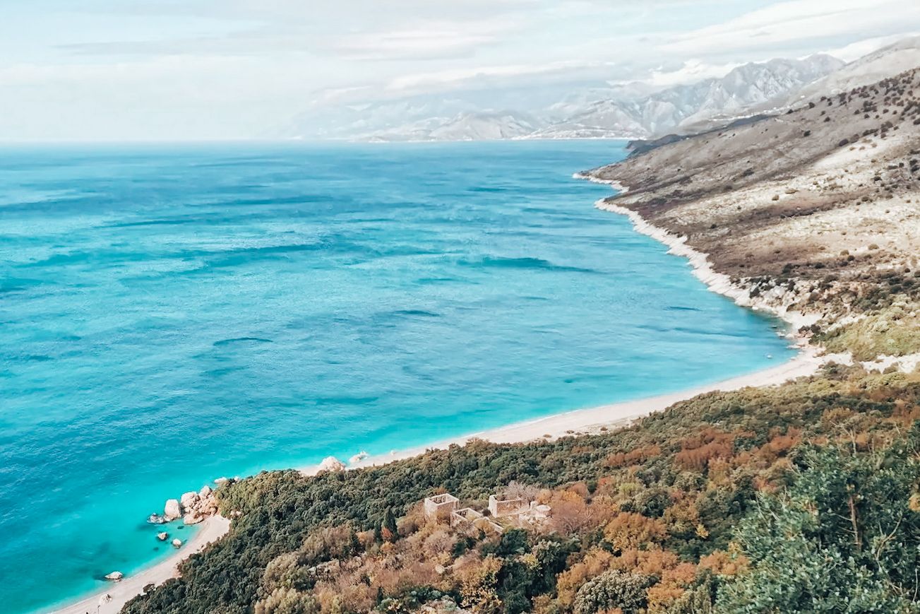 view of the Kroreza coastline in Albania showing a long stretch of bright turquoise sea meeting a mix of sandy shore and rocky cliffs. The water shifts in color near the coast, revealing shallow areas and submerged rock formations along the edge. This remote coastal area is known for its dramatic scenery and hidden sea caves along the Hidden Caves in Albania route.