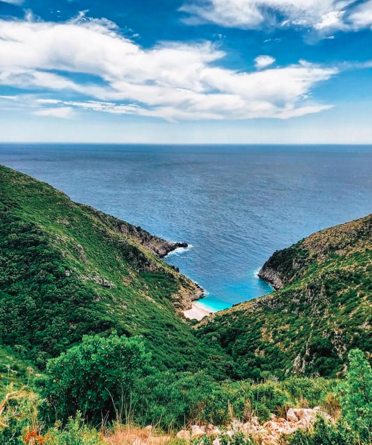 View of Gjiri i Dafines in Albania showing a quiet, sheltered bay with clear blue-green water enclosed by steep, rocky cliffs. The shoreline curves into a small cove with visible natural rock formations and a sense of isolation from nearby development. The rugged coastal geology hints at the Hidden Caves in Albania found throughout this stretch of coastline.
