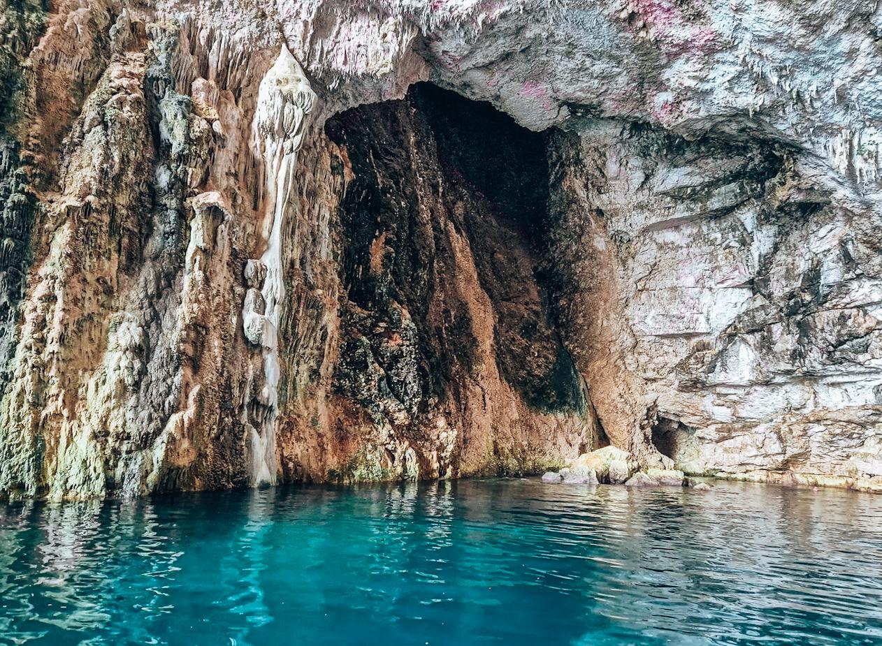 A sea cave entrance at Haxhi Alia Cave in Albania framed by towering limestone rock and deep blue water reflecting light inside the cavern. The opening reveals a partially hidden interior with rough stone textures shaped by waves over time. This striking coastal formation is one of the most iconic stops associated with Hidden Caves in Albania.