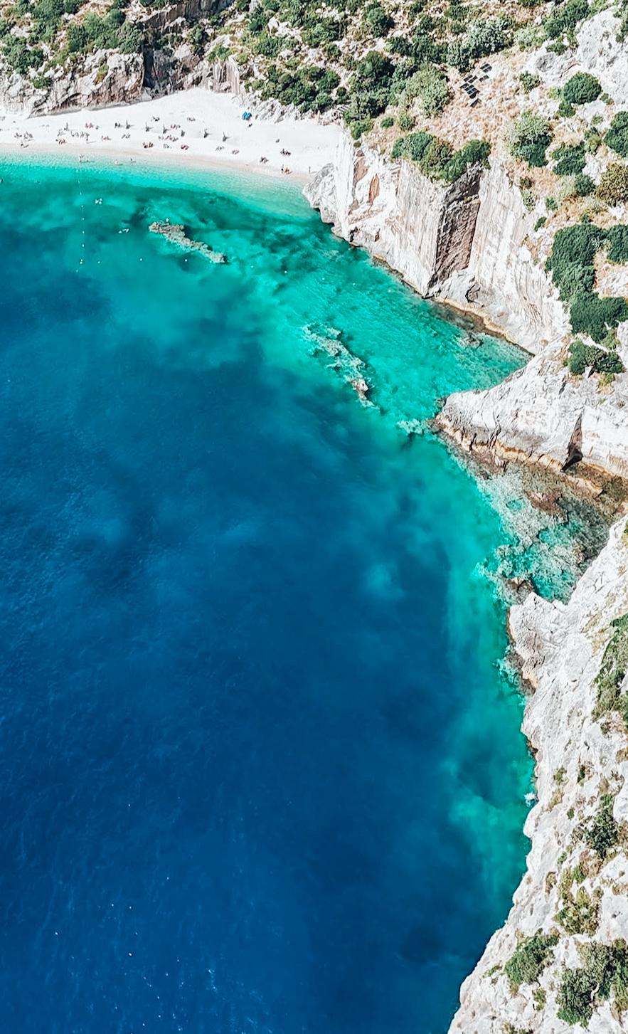 Aerial view of Grama Bay in Albania showing a secluded crescent-shaped beach surrounded by tall, rugged limestone cliffs and clear turquoise water. The coastline appears remote and largely untouched, with small coves and rock formations carved by the sea. The dramatic landscape reflects the hidden coastal caves and natural inlets associated with Hidden Caves in Albania.