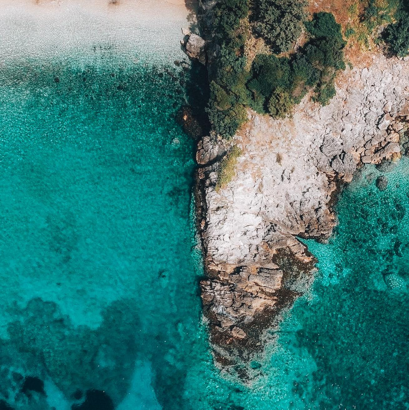 Aerial drone view of Qeefal Bay in Albania showing clear turquoise water meeting a rugged, rocky coastline with steep cliffs and small inlets. The scene captures the natural contrast between deep blue sea and pale limestone formations along the shore. This coastal landscape is part of the region known for Hidden Caves in Albania, where sea-carved formations and sheltered coves shape the shoreline.