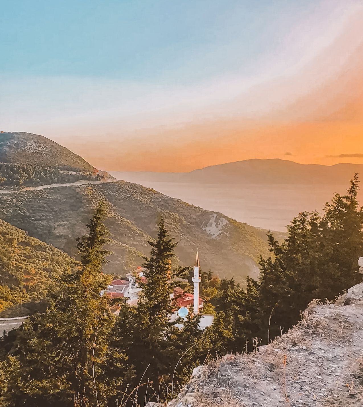 Kaninë Fortress overlooking the city of Vlorë at sunset with the sky glowing in soft orange and pink tones. The ancient stone walls frame a sweeping view of the city, coastline, and surrounding hills as daylight fades. A scenic elevated viewpoint capturing a peaceful evening atmosphere and one of the best sunset spots in Albania.