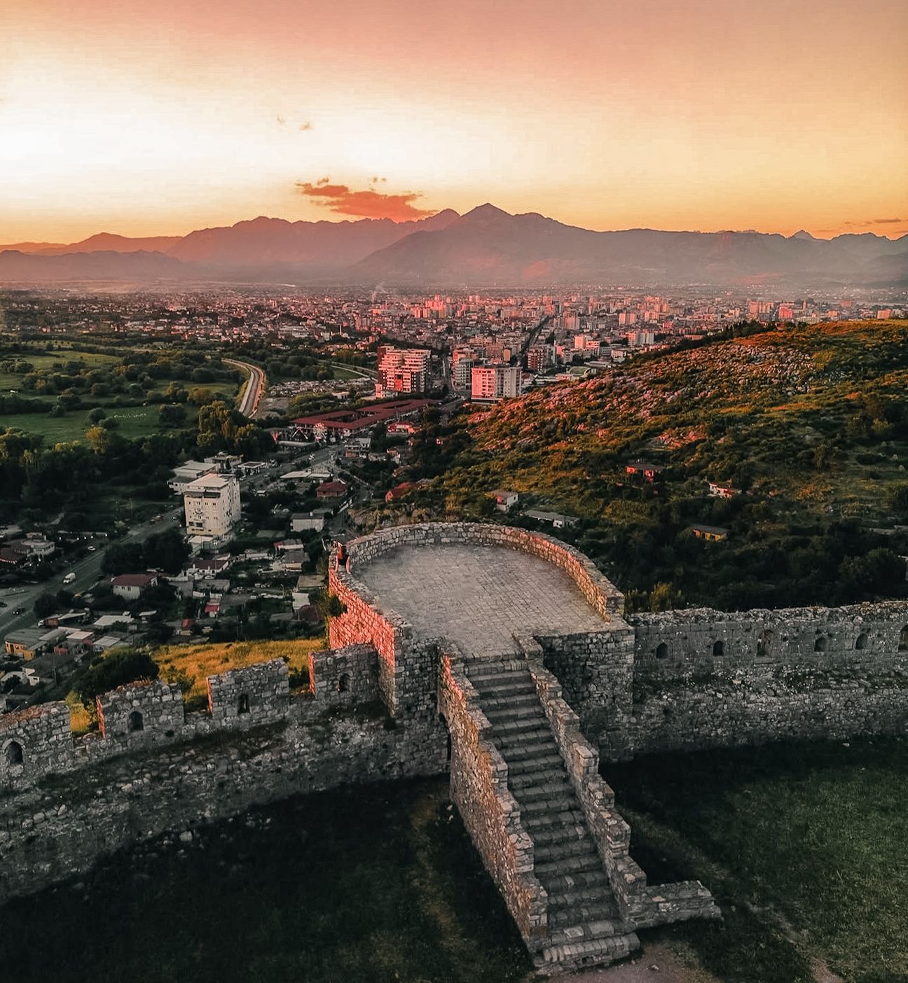 Rozafa Castle overlooking the city of Shkodër at sunset with warm golden light spreading across the landscape. The sun sets behind distant mountains, casting a soft glow over rooftops, the river, and surrounding hills. A peaceful elevated view capturing the historic fortress as one of the best sunset spots in Albania.