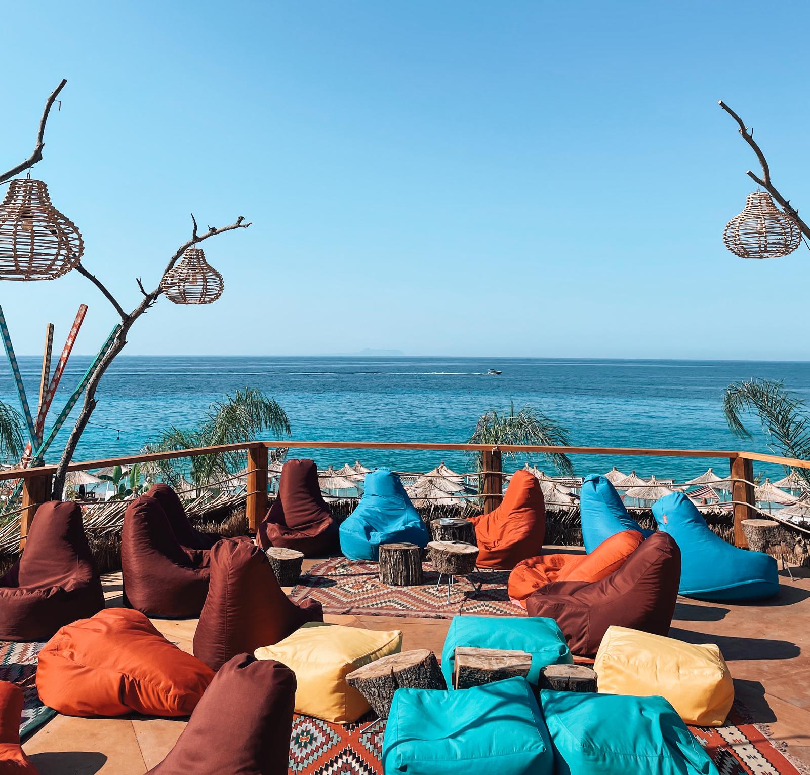 A relaxed beach bar in Dhermi with a laid-back coastal vibe, set close to the shoreline and surrounded by sun, sand, and summer atmosphere. The space appears designed for seaside drinks and socializing, capturing the contrast often seen in the drymades vs Dhermi stretch of coastline.