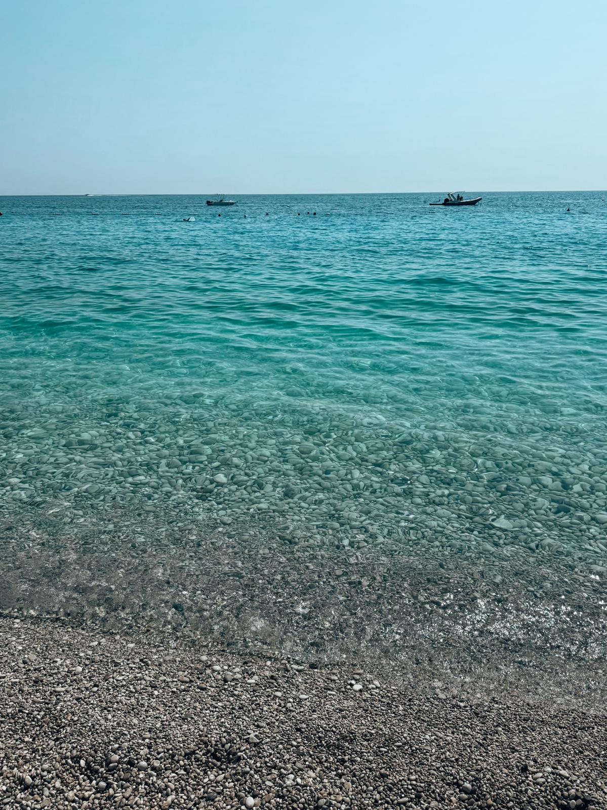 A calm stretch of sea along Drymades beach with clear blue water meeting a pebbled shoreline and open horizon. The scene captures the natural, less built-up coastal feel often contrasted in the drymades vs Dhermi comparison.