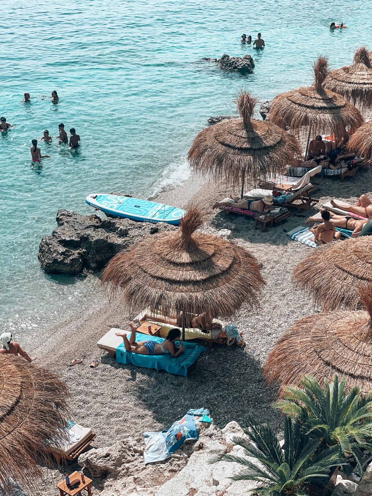 A wide beach in Dhermi with clear turquoise water gently meeting a shoreline of small stones and sand, framed by a relaxed coastal backdrop. The scene reflects the lively yet natural seaside atmosphere often discussed in the drymades vs Dhermi comparison.