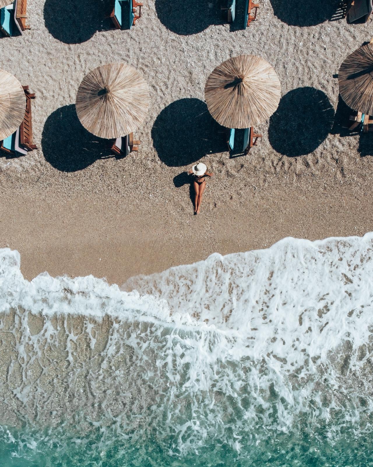 Aerial drone view over Drymades coastline showing blue sea meeting a rugged shore and scattered beach structures. The perspective highlights the contrast in the drymades vs Dhermi comparison emphasizing a more natural open coastline feel.