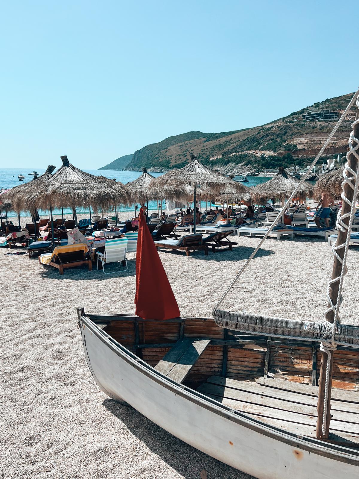 Bright summer scene at Jala Beach with clear turquoise water meeting a pebbled shoreline. Rows of sunbeds and umbrellas line the beach under strong sunlight, with a relaxed coastal atmosphere typical of an albanian riviera road trip stop along the Ionian coast.