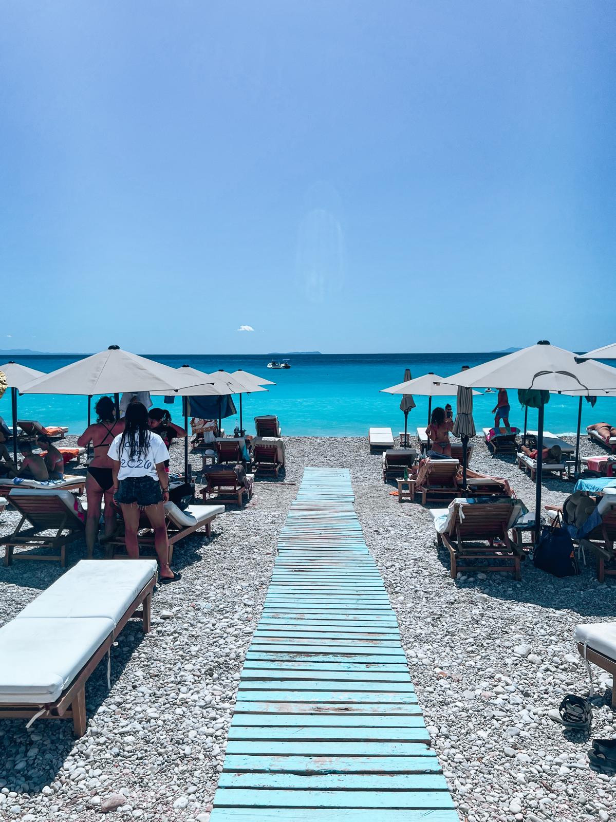Calm shoreline at Qeparo Beach with clear blue water gently meeting a pebbled and sandy coast. A quiet coastal village sits above the beach with hillside homes overlooking the sea, evoking a peaceful stop along an albanian riviera road trip.