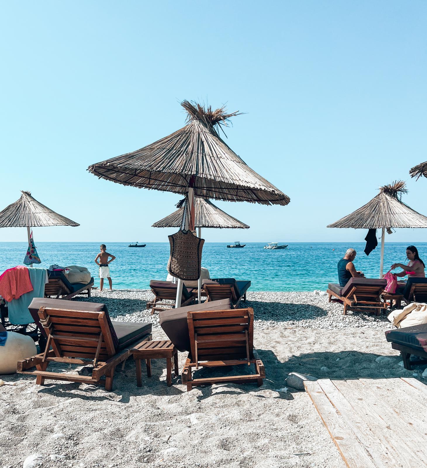 Bright coastal scene along the South Riviera with calm blue sea stretching toward the horizon under strong sunlight. Sunbeds and umbrellas are arranged along the shoreline, creating a relaxed beach atmosphere typical of an albanian riviera road trip stop.