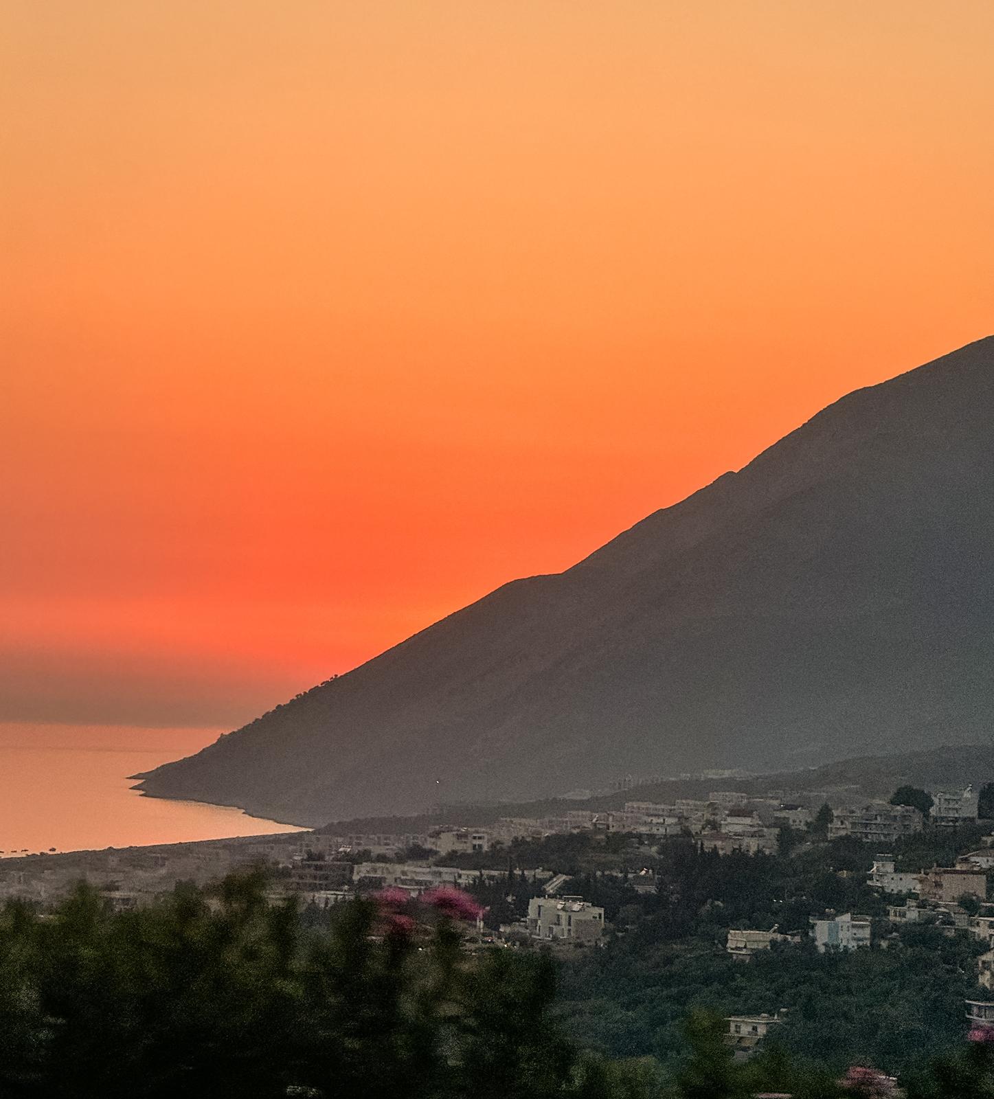 Alt text: Golden sunset over the coastal village of Dhermi seen through a car window on a winding seaside road. The sky glows orange and pink as cliffs and the Ionian coastline fade into silhouette capturing a moment on an albanian riviera road trip.