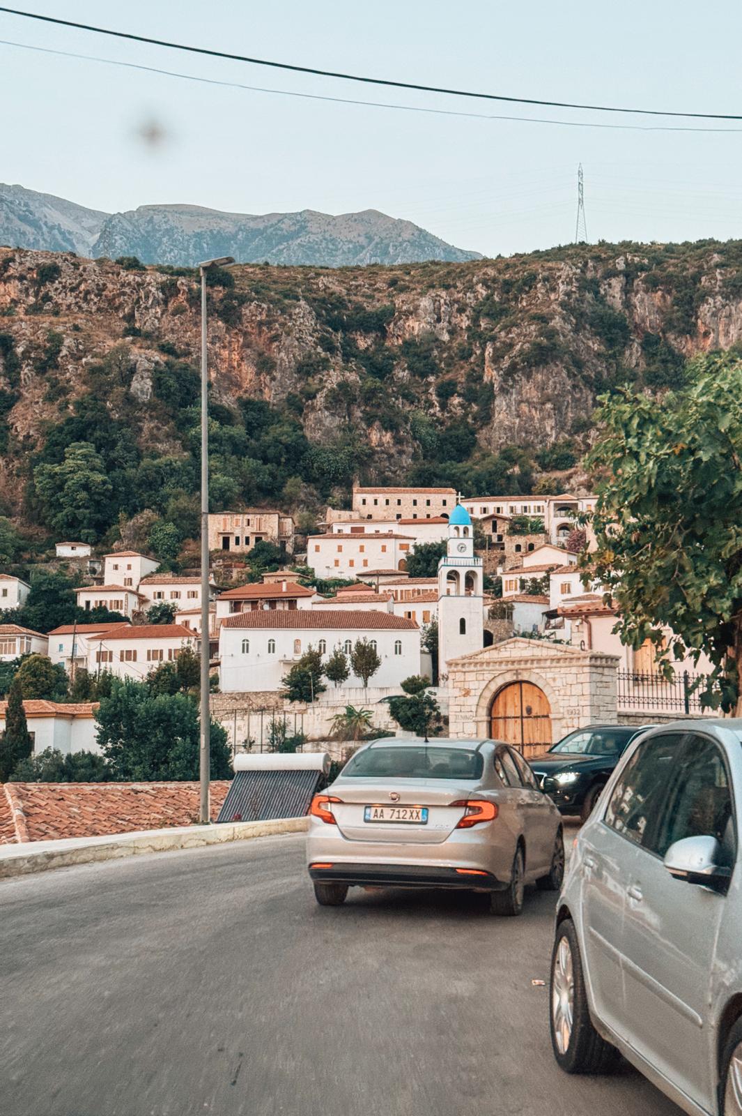 View of Drymades village along the Albanian coast seen from a moving car, with clustered houses and greenery stretching toward the sea. The coastline and hills blend into the background under bright daylight, capturing a scenic moment on an albanian riviera road trip.