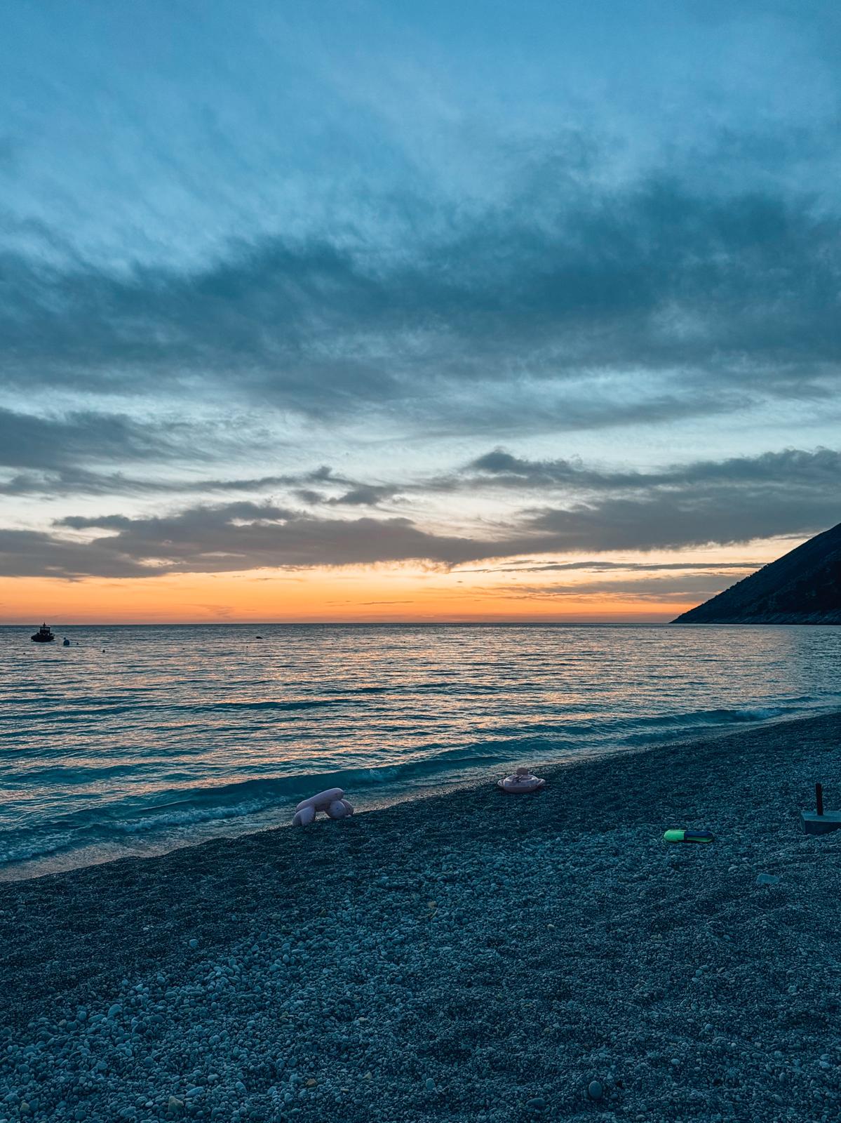 Warm sunset over Palasa along the Albanian coast with golden light reflecting across the sea and surrounding shoreline. The sky shifts into soft orange and pink tones as the landscape fades into silhouette, capturing a scenic moment on an albanian riviera road trip.