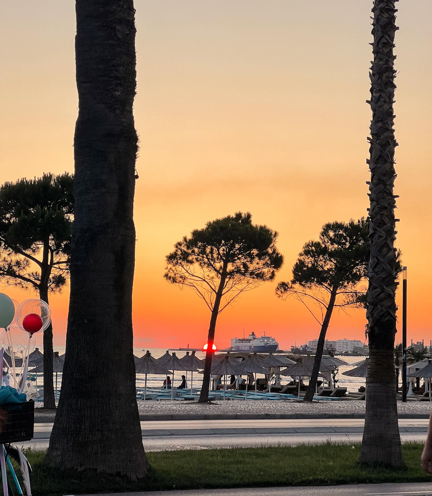 Sunset view of Vlora seen from inside a car with the coastline stretching along the horizon. The sky glows in warm orange and pink tones over the sea and city edge, capturing a moving moment during an albanian riviera road trip.