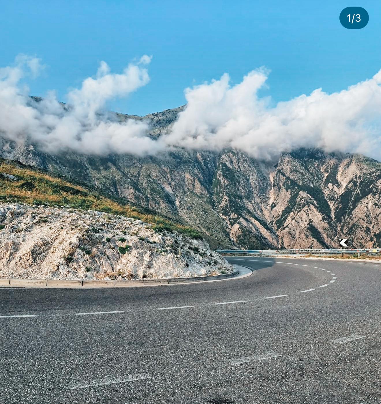 Mountain pass view at Llogara Pass with dramatic green peaks rising above winding roads and forested slopes. Clouds drift over the high ridge as the coastline becomes visible in the distance, marking a scenic transition point on an albanian riviera road trip.
