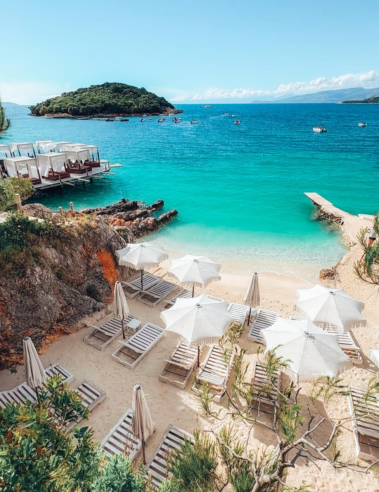 Crystal clear turquoise waters at Ksamil Beach with small islands visible just offshore under bright summer sunlight. The shoreline is lined with sand and beachgoers relaxing in a vibrant coastal setting that feels like a highlight stop on an albanian riviera road trip.
