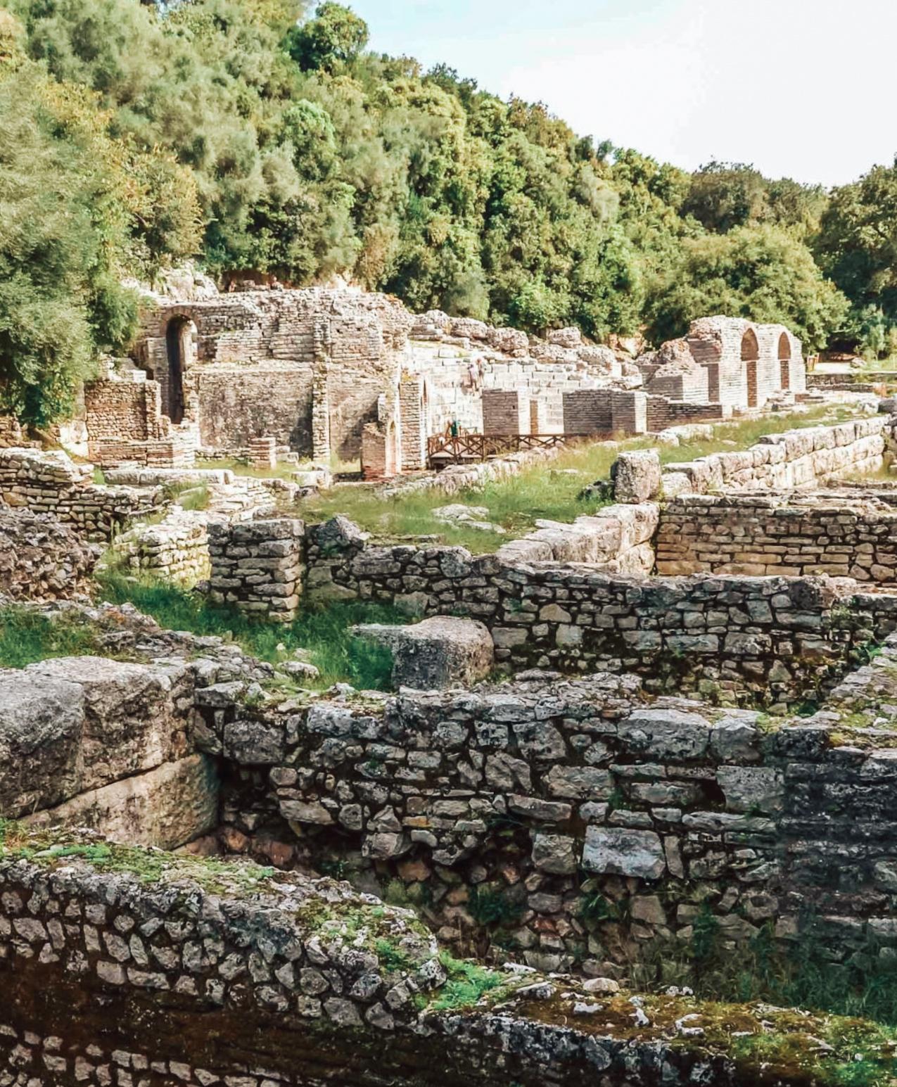 Ancient ruins of Butrint surrounded by lush greenery and calm water, with stone structures and pathways hinting at its long archaeological history. The peaceful natural setting contrasts with the historic remains, making it a memorable cultural stop on an albanian riviera road trip.
