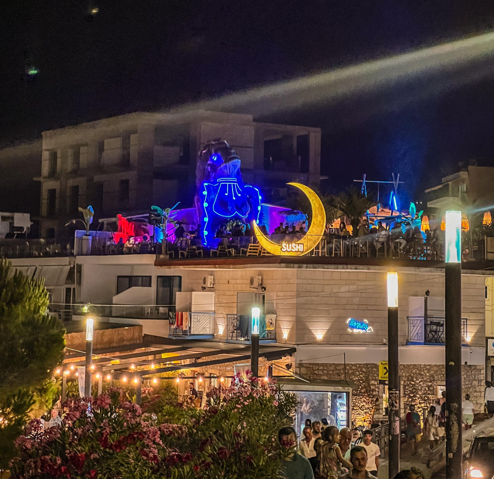 Night view of Himara city center with warm streetlights illuminating the buildings and streets. The town appears calm after dark, with a soft glow reflecting off nearby surfaces and creating a quiet coastal evening atmosphere.