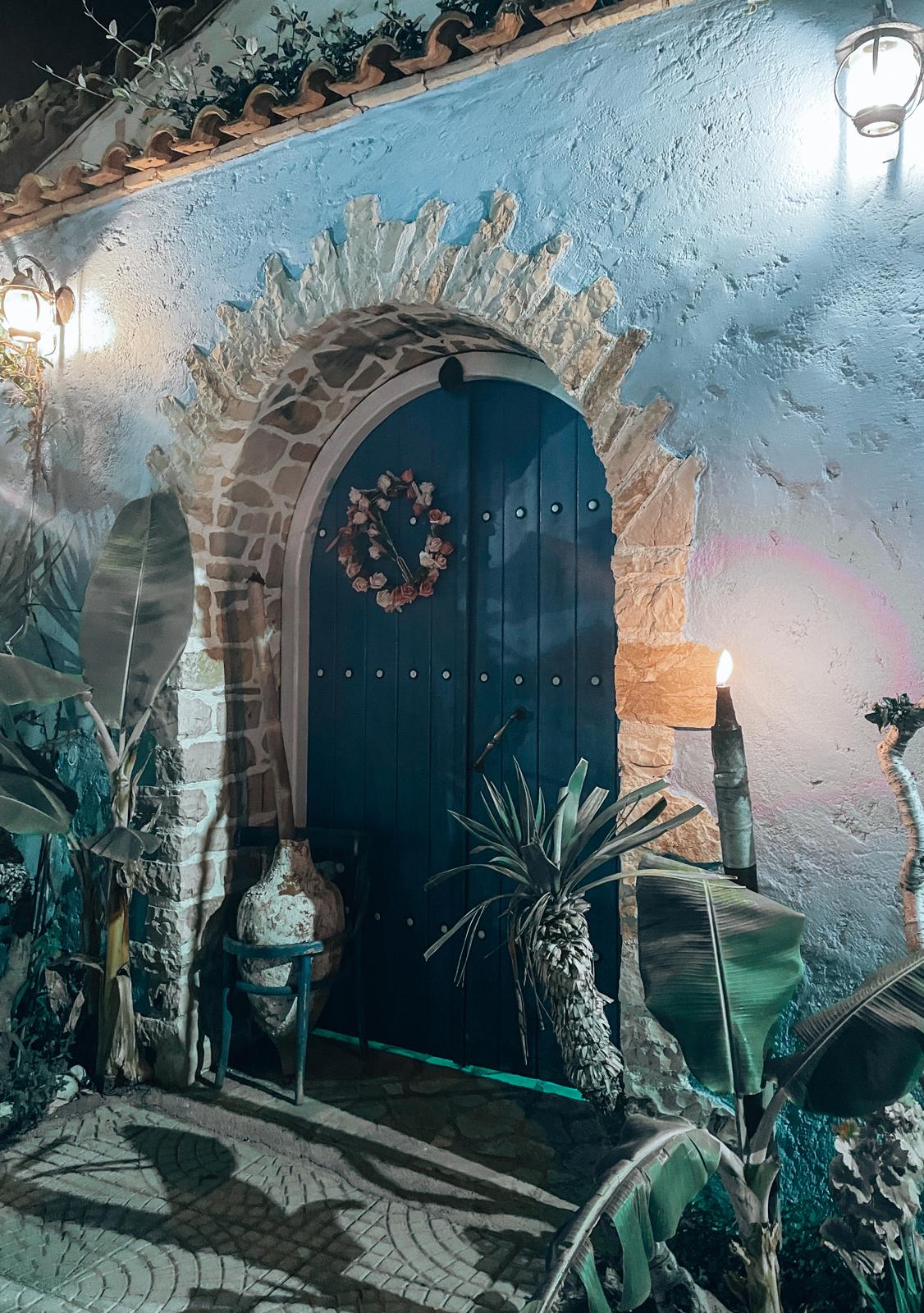 Decorative wooden door set into a stone building facade in Himara city center, featuring traditional design details and textured masonry around it. The image highlights architectural character in the old town area, reflecting the historic coastal charm of Himara.