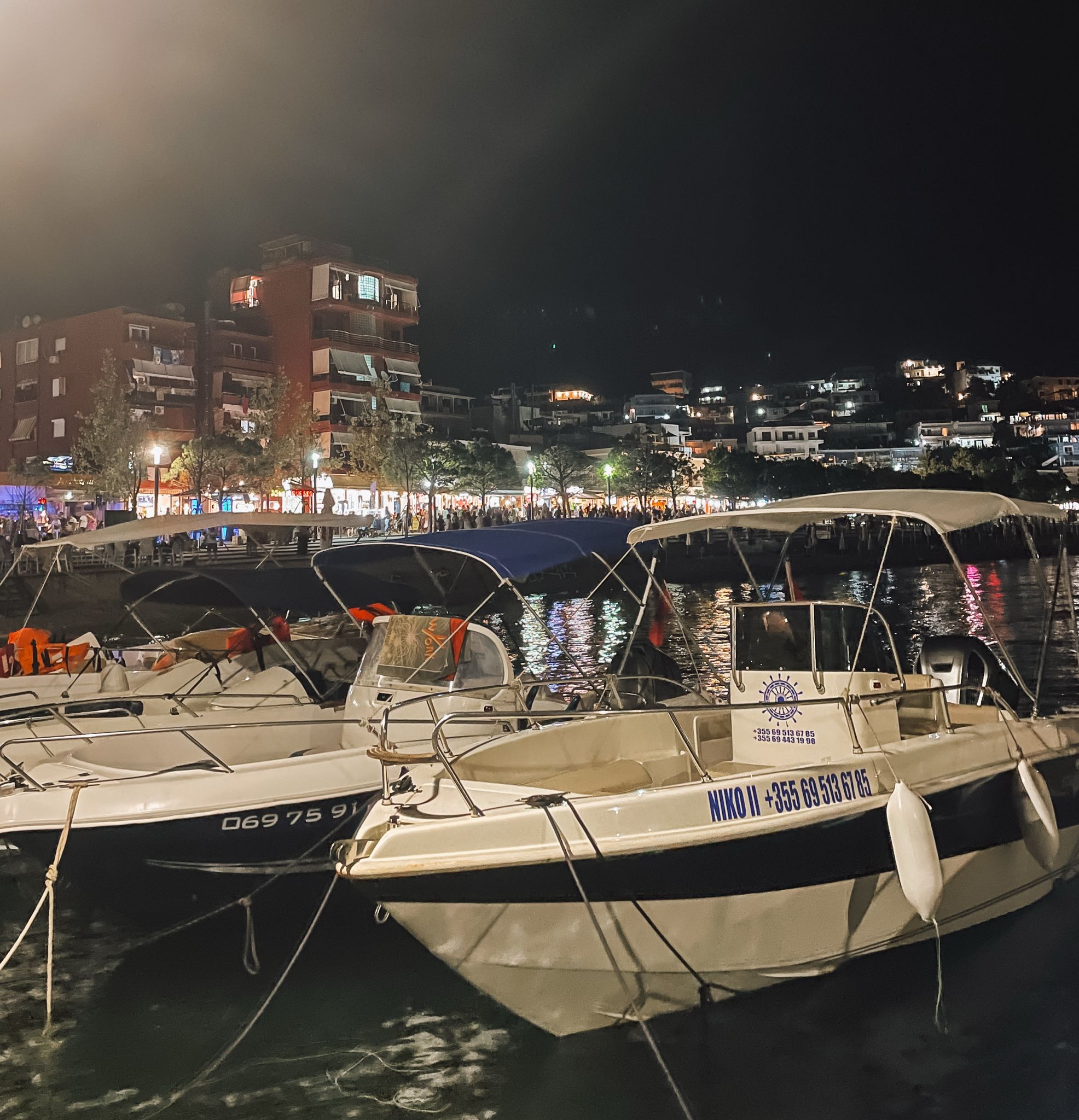Himara port with small fishing boats and yachts docked along calm blue water, surrounded by the coastal town and nearby hills. The harbor area reflects the relaxed seaside atmosphere and highlights one of the scenic locations near where to stay in Himara.