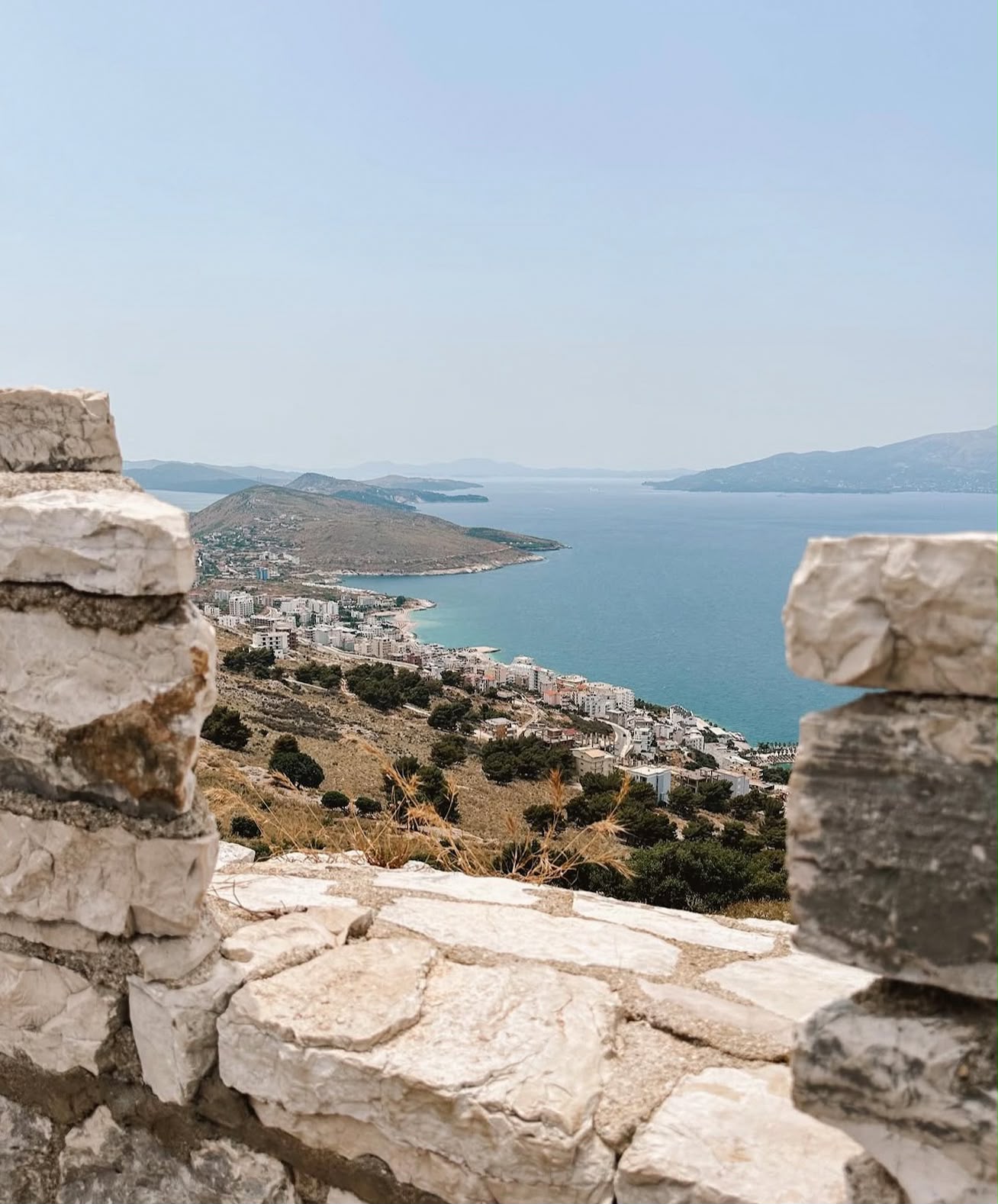 Stone walls of Lëkurësi Castle sitting on a hill overlooking Sarandë and the Ionian Sea. The elevated viewpoint shows sweeping coastal views with the city and shoreline stretching below.