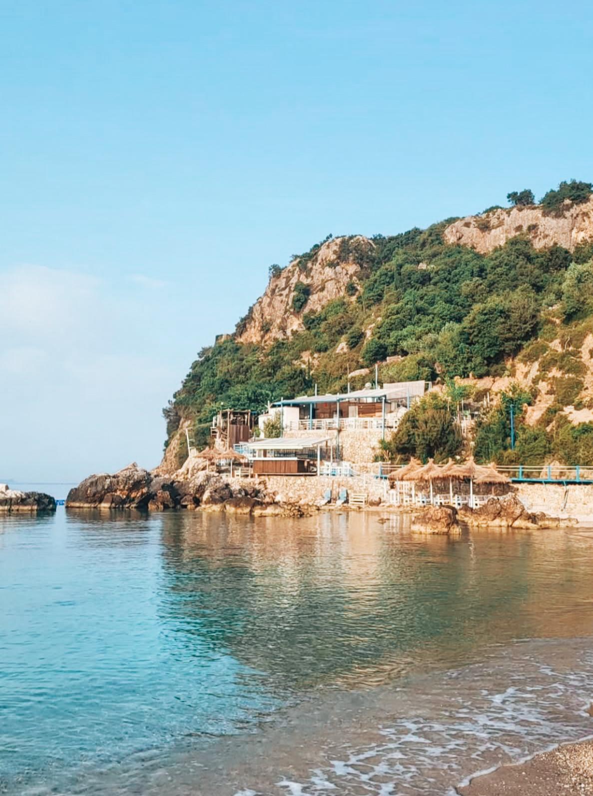 Wide view of Livadhi Beach with clear turquoise water, a long stretch of white pebbles, and green hills surrounding the bay on a sunny day.