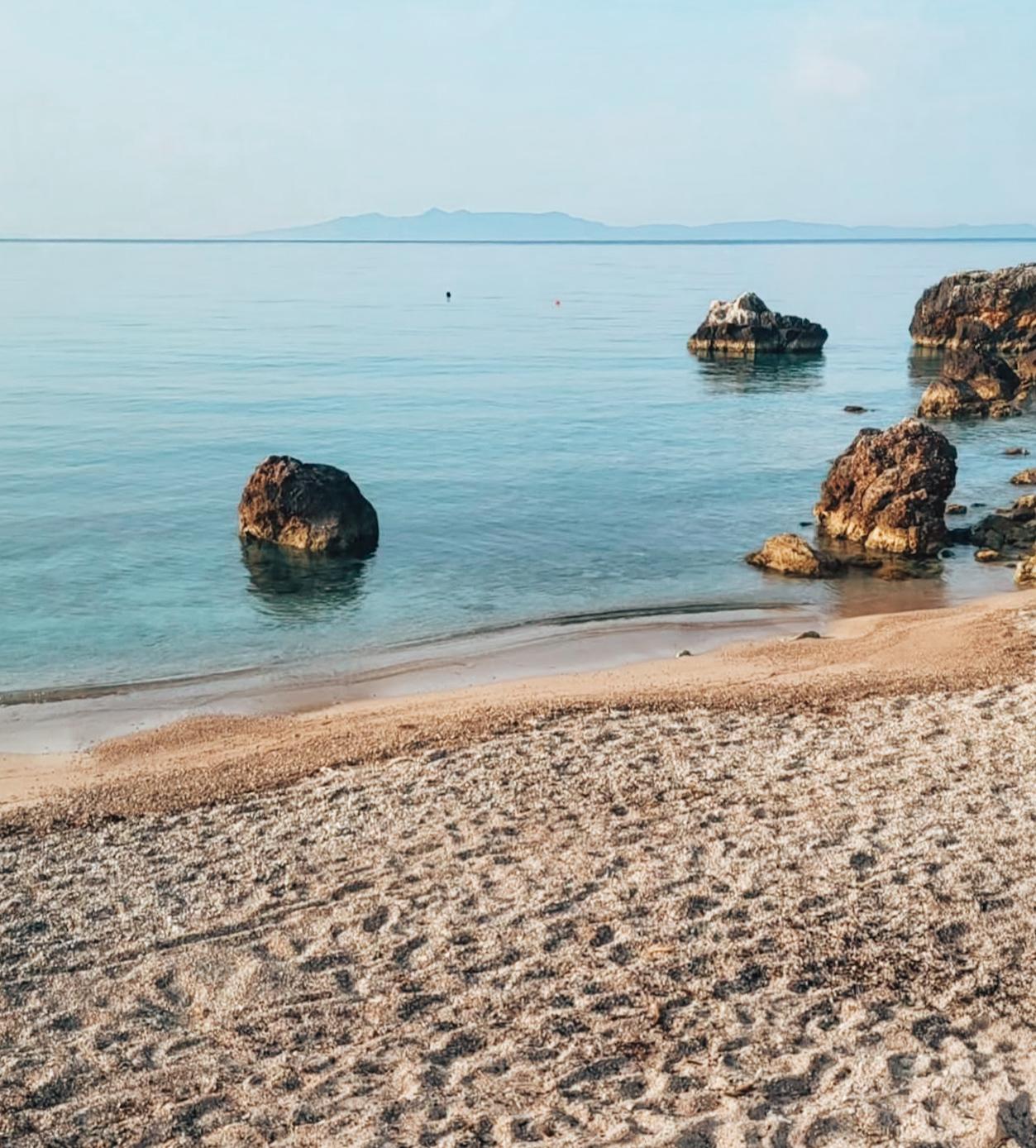 Crystal clear water laps against the pebbled shoreline at Livadhi Beach as visitors swim and relax along the coast.