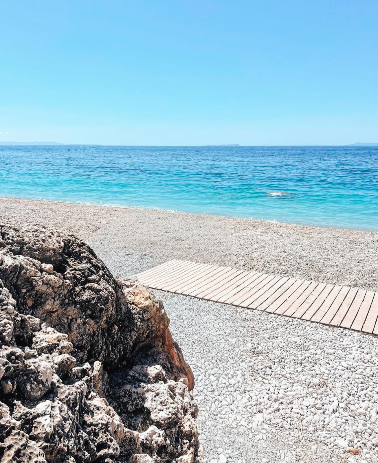 Panoramic view of Livadhi Beach showing the calm blue sea, beach umbrellas, and the mountainous coastline in the background.