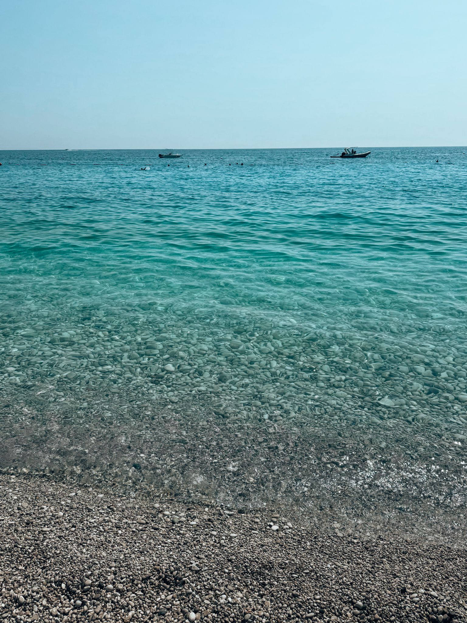 Livadhi Beach near Himara with a long stretch of light-colored pebbles and clear blue sea gently meeting the shore, backed by rolling green hills. The spacious, relaxed coastline highlights why this area is popular for travelers choosing where to stay in Himara for a quieter seaside escape.